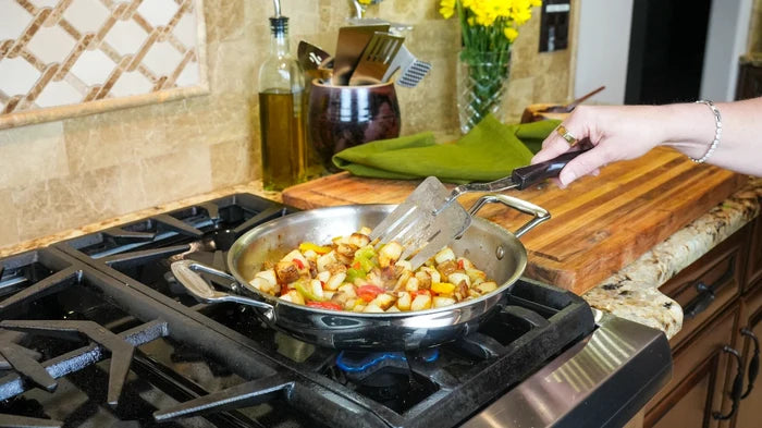 Hand stirring diced potatoes and peppers in a stainless steel 10-inch fry pan on a gas stove in a home kitchen
