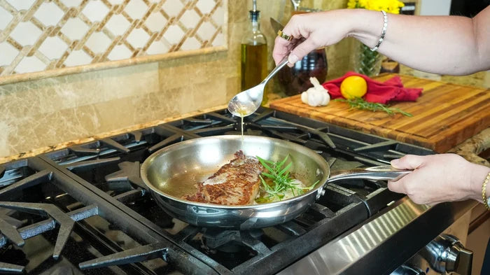 Stainless steel 10-inch fry pan searing a steak on a gas stove as a hand spoons pan sauce over the meat
