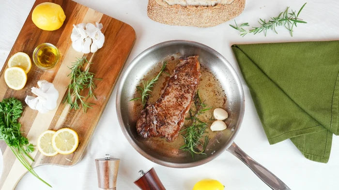 Top-down view of a 10-inch stainless steel fry pan searing a steak with garlic, rosemary, and lemon on a white tabletop