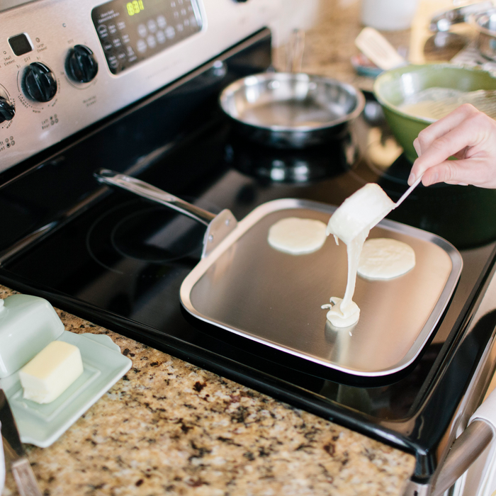 11-inch square stainless steel griddle on a stovetop with a hand pouring pancake batter and three small pancakes cooking