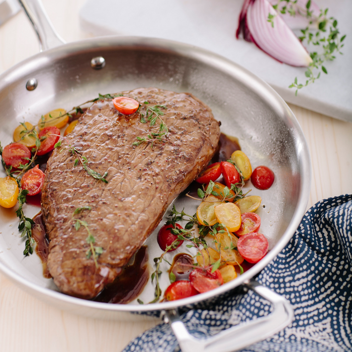 Angled top-down view of 11.5 inch stainless steel fry pan showcasing brushed interior and ergonomic stay-cool handle with seared steak and cherry tomatoes on a light wood surface