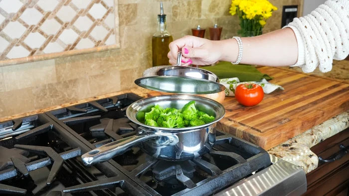 Hand lifting lid of 1.75 quart saucepan to reveal broccoli on a gas stove with cutting board and tomato