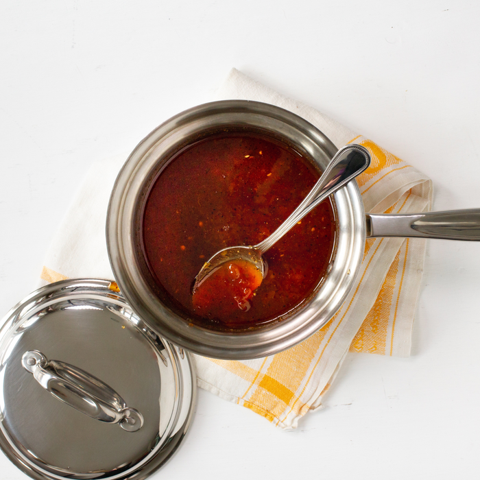 Stainless-steel saucepan with red tomato sauce and spoon, lid beside, on folded yellow-striped dish towel