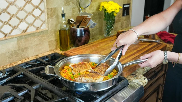 Hands using tongs to lift chicken in a stainless steel saute pan with rice on a gas stove, cutting board and flowers behind