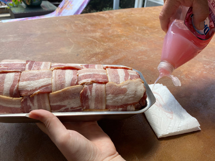 Side view of stainless steel mini jelly roll pan with bacon-wrapped meatloaf, showing rolled edge as hand pours pink liquid