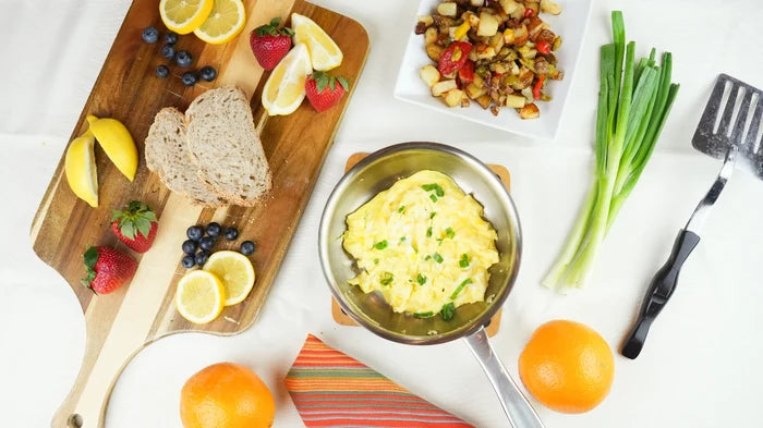 Top view of 7-inch stainless steel fry pan holding scrambled eggs and scallions on white table with bread, fruit and spatula