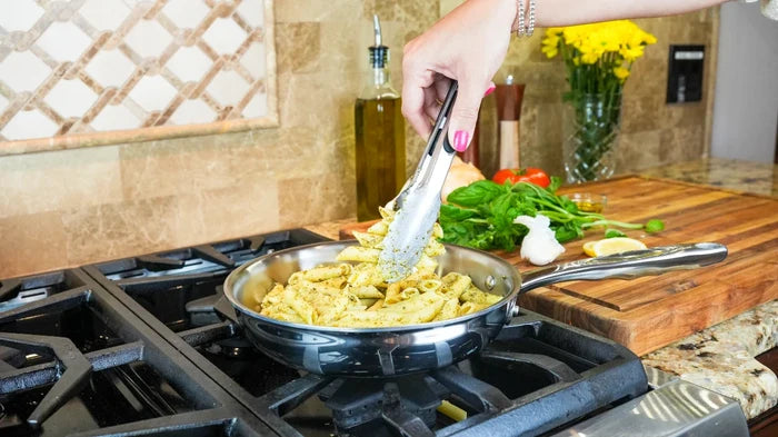 Person using tongs to serve pasta from an 8.5-inch stainless steel fry pan on a gas stove with cutting board and vegetables