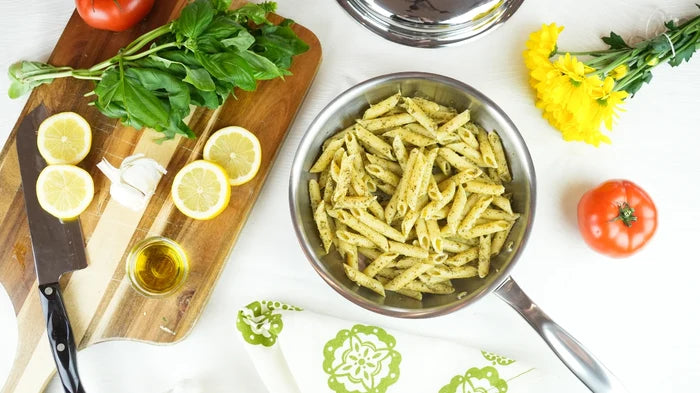 8.5-inch stainless steel fry pan holding pesto penne beside cutting board with lemon, basil, and tomato on white surface