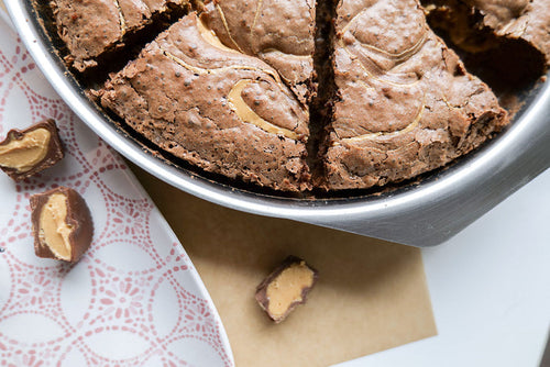 Top-down view of 9-inch round brownie pan showing brushed stainless steel rim, integrated tab handle, and peanut-butter swirl brownies on white background