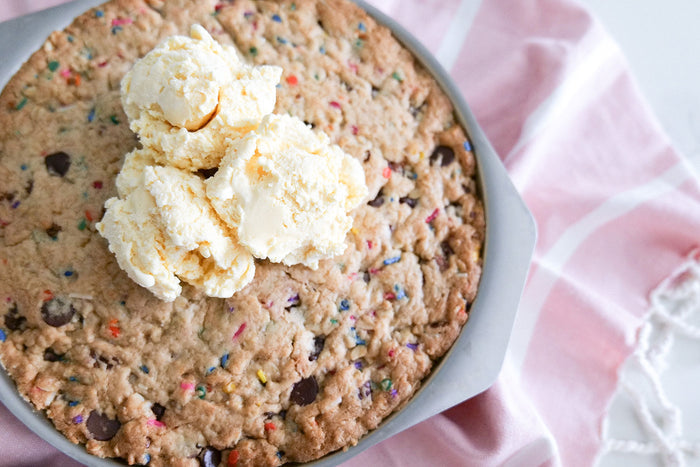 Top-down view of 9-inch round stainless steel baking pan showcasing a cookie cake topped with scoops of vanilla ice cream on a pink cloth background