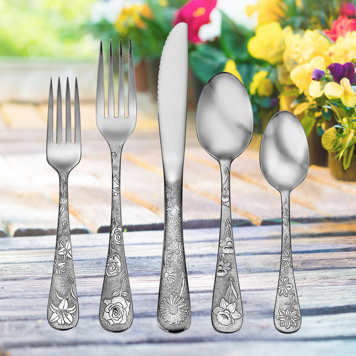 Five-piece floral-engraved flatware (2 forks, knife, tablespoon, teaspoon) on wooden table with blurred flowers.