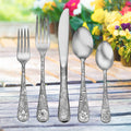 Five-piece floral-engraved flatware (2 forks, knife, tablespoon, teaspoon) on wooden table with blurred flowers.