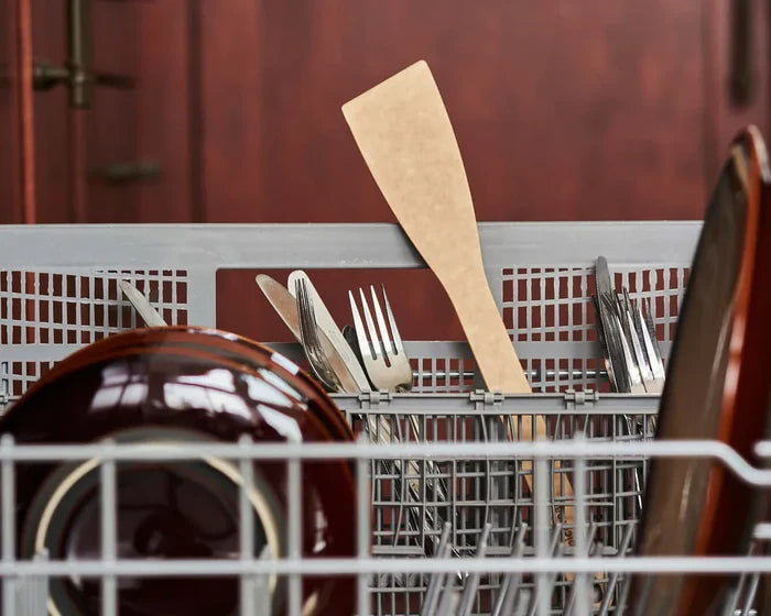 Close-up of Angled Turner standing upright in a dishwasher cutlery basket, showing angled flat blade and natural wood handle among dishes