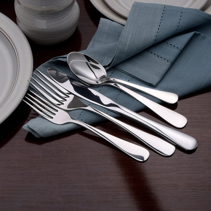Polished stainless-steel forks, spoons, and knives arranged on a blue cloth napkin beside plates on a dark wood table.