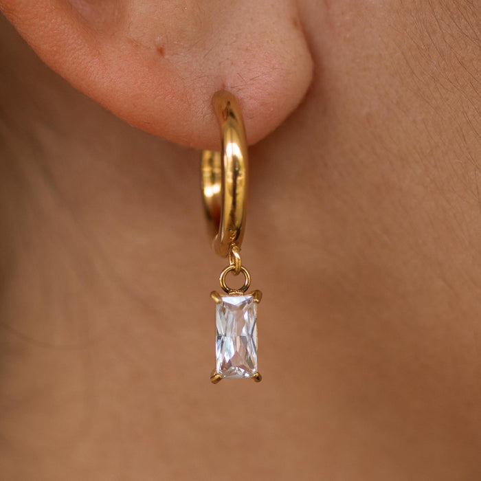 A gold huggie earring with a dangling baguette-cut stone, displayed on a person's ear.
