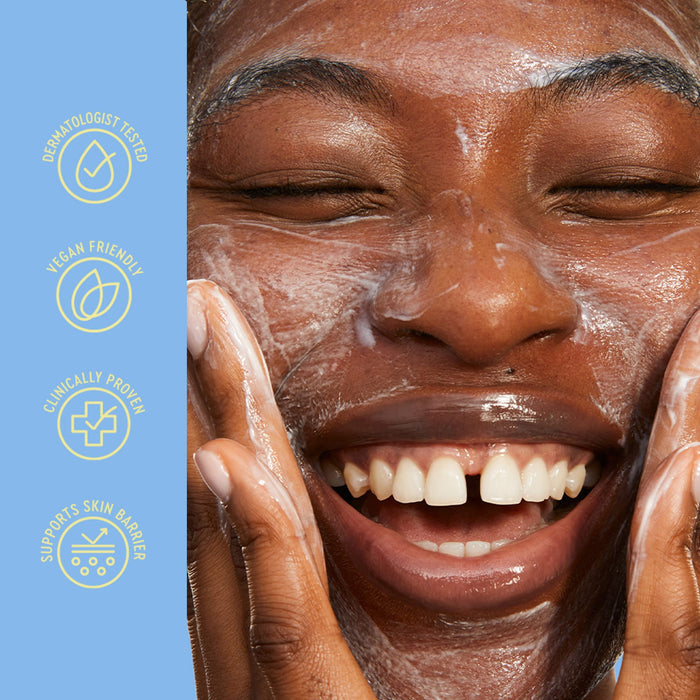 Close-up of a smiling person with white cleanser or soap on their face, touching their cheeks, with a light blue vertical banner on the left containing yellow icons and text reading 'Dermatologist Tested', 'Vegan Friendly', 'Clinically Proven', and 'Supports Skin Barrier'.