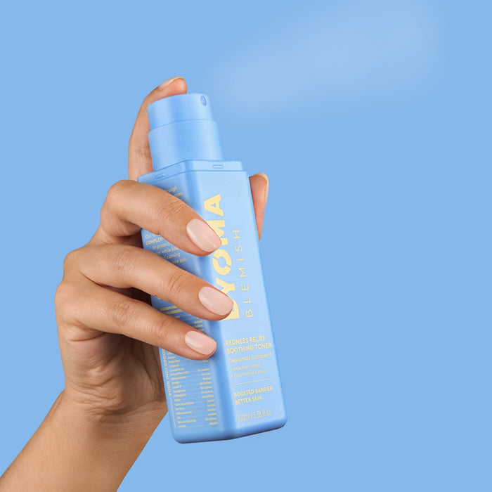 A hand with light pink manicured nails holds a light blue spray bottle of Icomate hair growth serum against a solid light blue background.