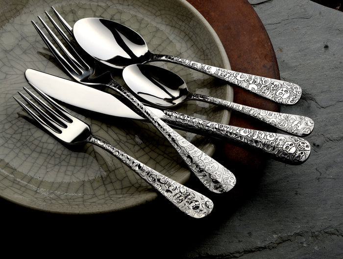 Angled view of Calavera flatware showing skull-motif handles on forks, spoons, and knives on a ceramic plate