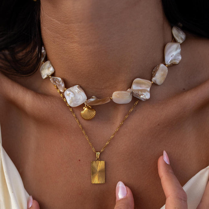 close up of a womans neck wearing a gold twist necklace with a rectangle sunburst pendant and a baroque pearl necklace with a gold shell pendant