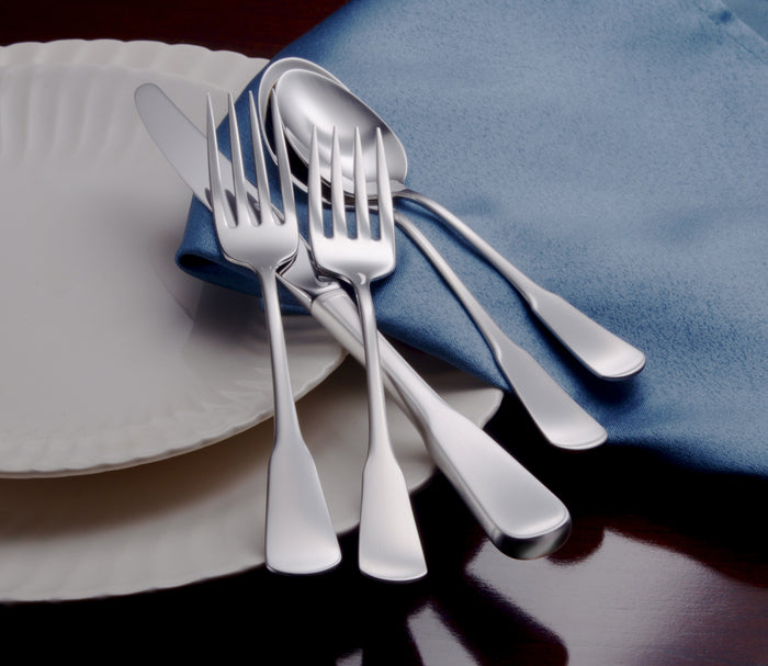 Angled view of Candra 45-piece flatware set showcasing brushed satin handles and mirror-finish bowls on a blue napkin beside a white plate