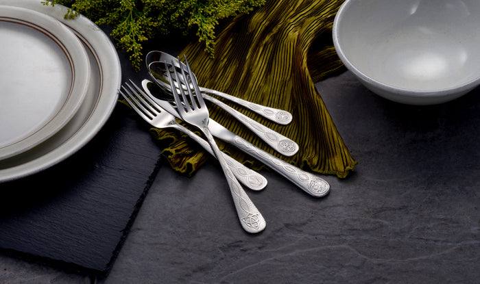 Set of ornate silver forks and spoons arranged on a dark surface with stacked white plates, a white bowl, and a green textured cloth.