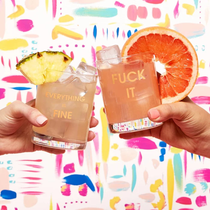 Two hands clink Chez Gagné More Rocks Glasses—one gold-foil stamped “EVERYTHING IS FINE,” the other “FUCK IT”—against a vibrant, abstract background with playful brushstrokes and dots. Brand: Chez Gagné.
