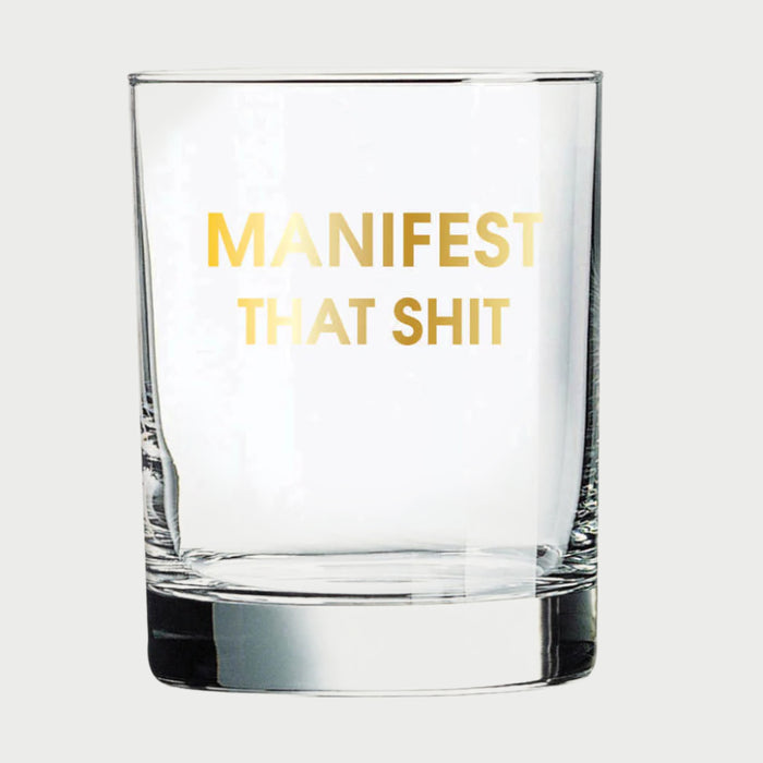 The Chez Gagné More Rocks Glasses by Chez Gagné feature a gold foil "MANIFEST THAT SHIT" imprint on a clear glass, shown against a simple light background.