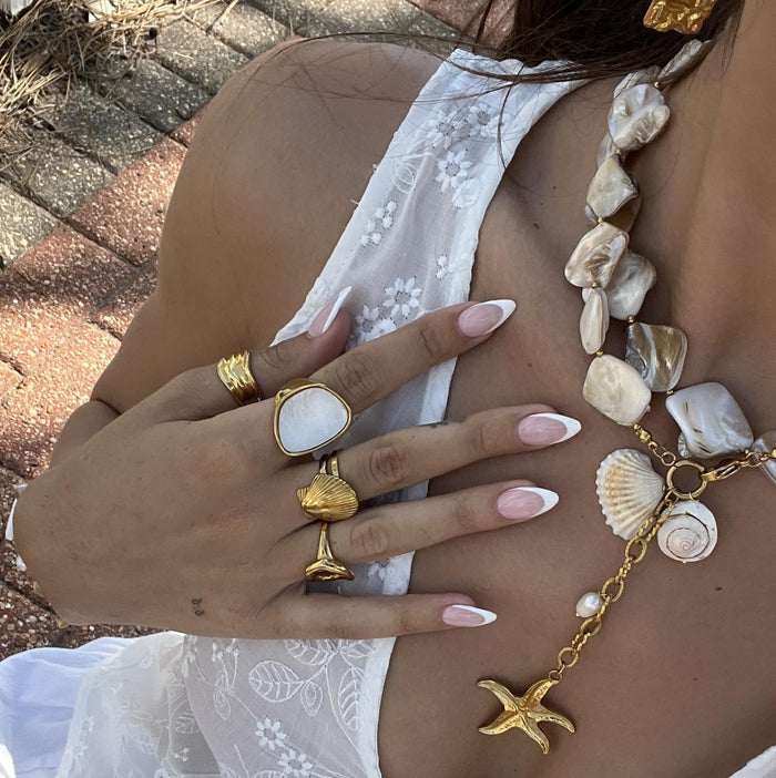 Close up of a womans neck and hand wearing chunky vacation jewelry featuring shells, charms, baroque pearls and waterproof gold designs
