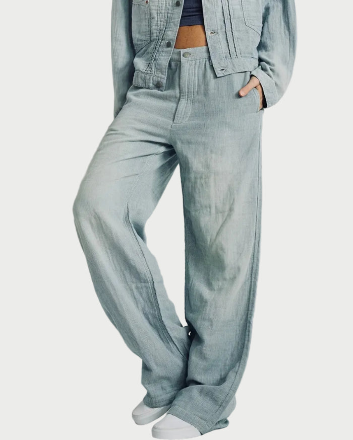A person wearing light-wash wide-leg denim pants with a matching cropped denim jacket and white sneakers, standing against a plain light gray studio background.