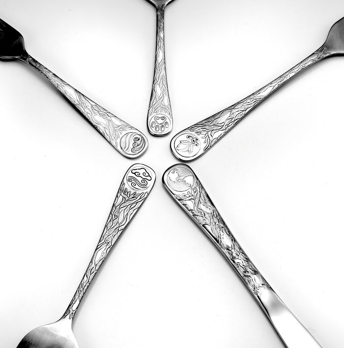 Top view of Earth flatware spoons showcasing engraved sun, moon, wave, and fish motifs on polished handles against a white background