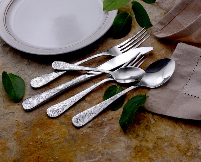 Top view of Earth pattern flatware showing engraved planet motifs on handles with white plate, green leaves, and tan napkin