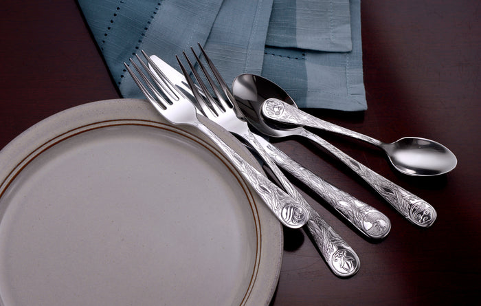 Angled top view of Earth pattern flatware showing engraved sun, moon, and wave motifs on handles with white plate, gold rim and blue napkin on dark wood table