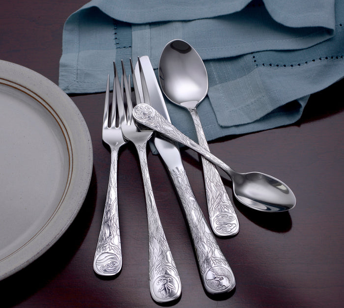 Top view of Earth flatware showing engraved nature motifs on handles with blue napkin and white plate on dark wood table