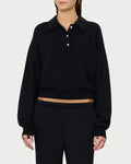 A person wearing the Éterne Cropped Polo Sweatshirt by Eterne—a relaxed fit, black polo sweatshirt featuring three buttons and a slightly cropped length that reveals a hint of midriff—is paired with matching black pants and stands against a plain light background.