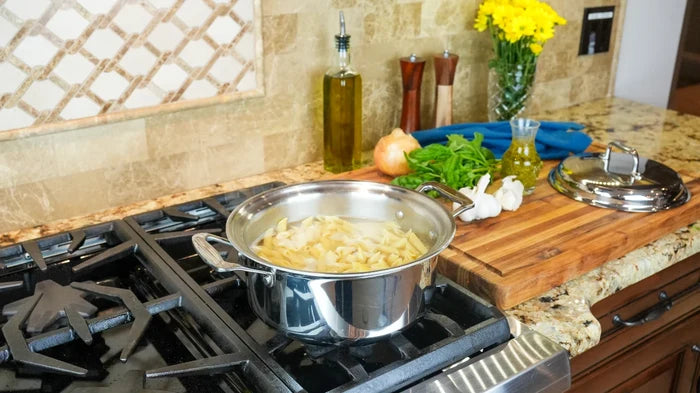 6-quart stainless steel stockpot simmering pasta on a gas stove in a kitchen with a cutting board, garlic, onion, and olive oil bottle