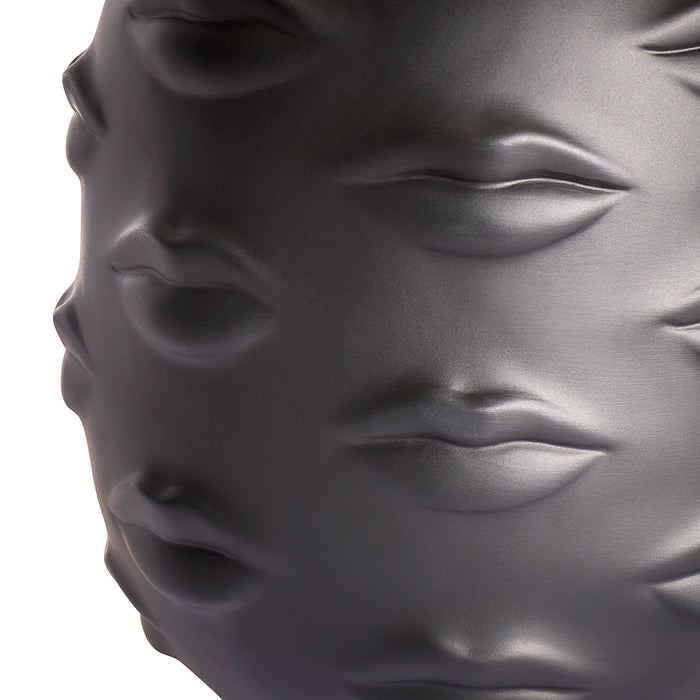 Close-up of a glossy dark gray sculptural object featuring embossed lips and eyes, photographed against a white background.