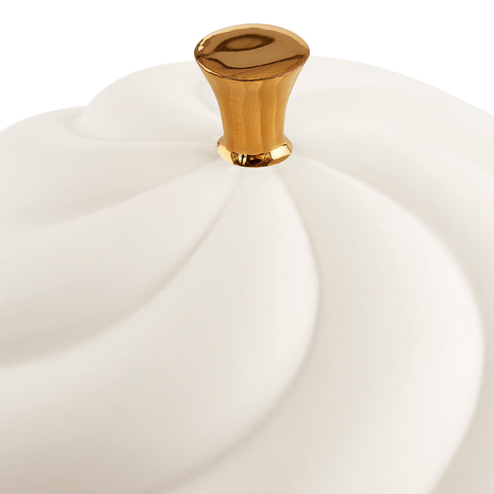 A close-up of a white ceramic container lid with a sculpted, swirled design and a polished gold knob on a white background.