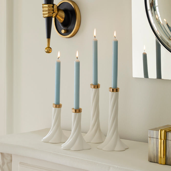Four white twisted candlestick holders with gold accents holding light blue taper candles, styled on a white mantelpiece against a cream wall with a wall sconce and mirror visible.