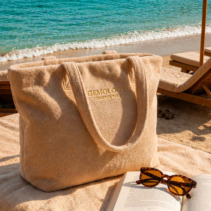 Grand sac de plage beige Gemology posé sur le sable avec livre et lunettes de soleil, vue mer