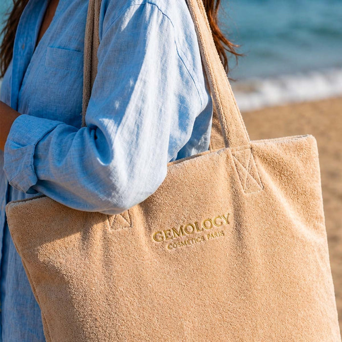 Femme portant le sac de plage beige Gemology à l'épaule au bord de la mer, logo brodé doré visible