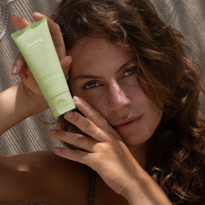 A woman with curly hair holds a tube of Tropic Hand Therapy softening superfood cream beside her face, smiling gently in warm sunlight.