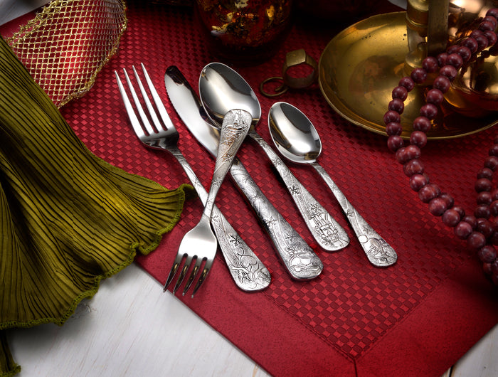 Engraved silver flatware (two forks, three spoons) arranged on red placemat with folded green napkin, brass candlestick and beaded necklace