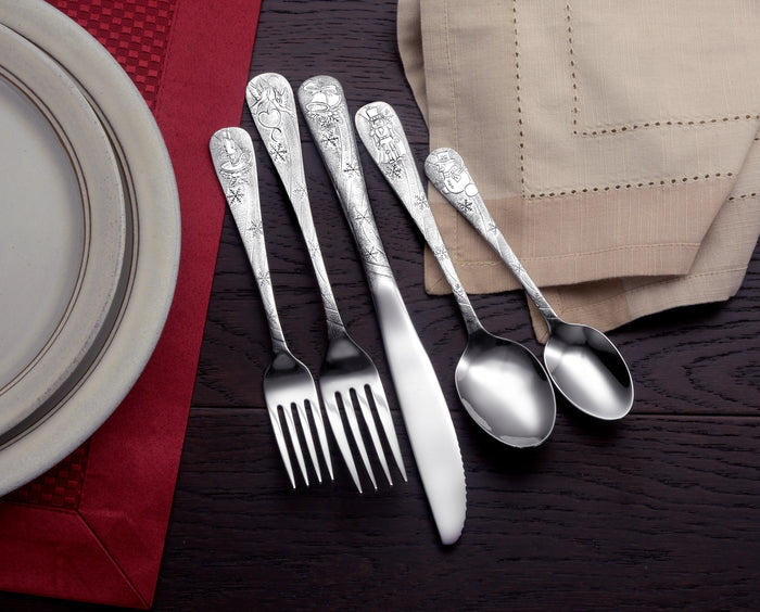 Ornate silver flatware, two forks, a knife and two spoons, arranged on dark wood beside a plate and folded napkins.