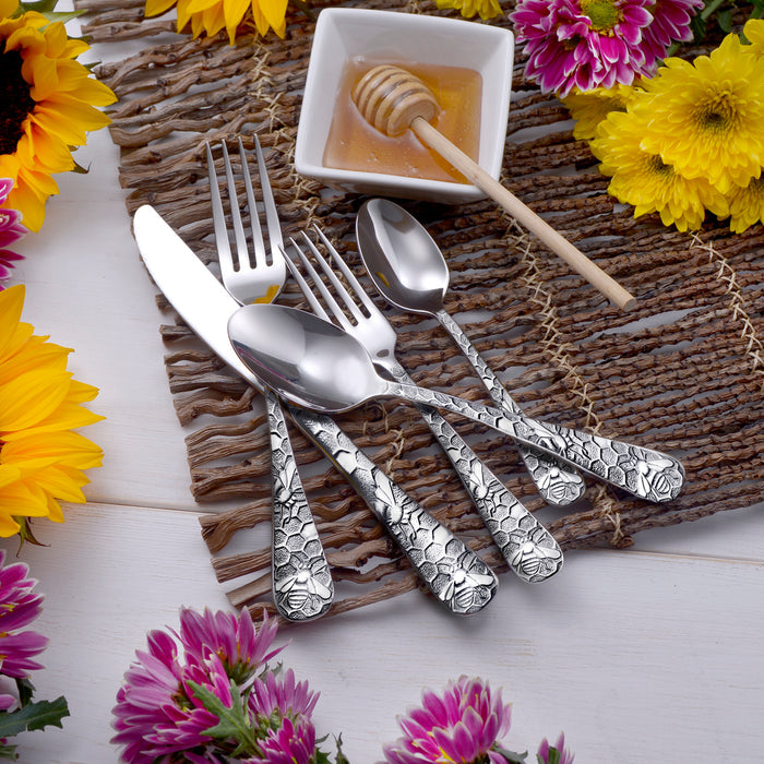 Angled view of Honey Bee 45-piece set flatware showing honeycomb bee-patterned handles on woven placemat with honey dipper