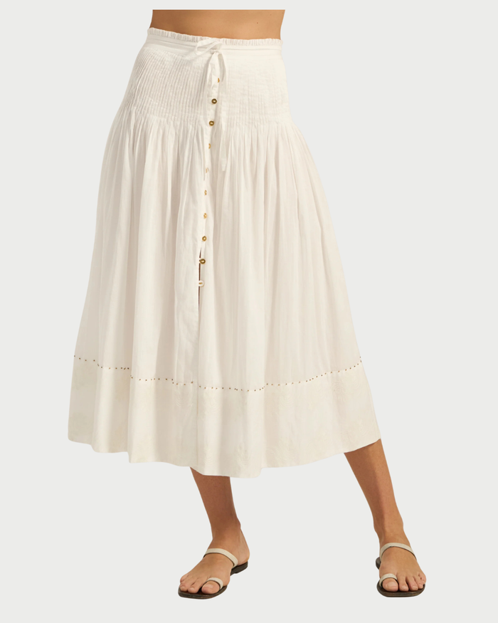 A woman wears the Ilio Nema Diana Skirt, crafted from organic white voile with subtle stitching, a tie waist, and button-front details. Its relaxed fit and flowy silhouette from Ilio Nema pair effortlessly with flat sandals for elegant ease.