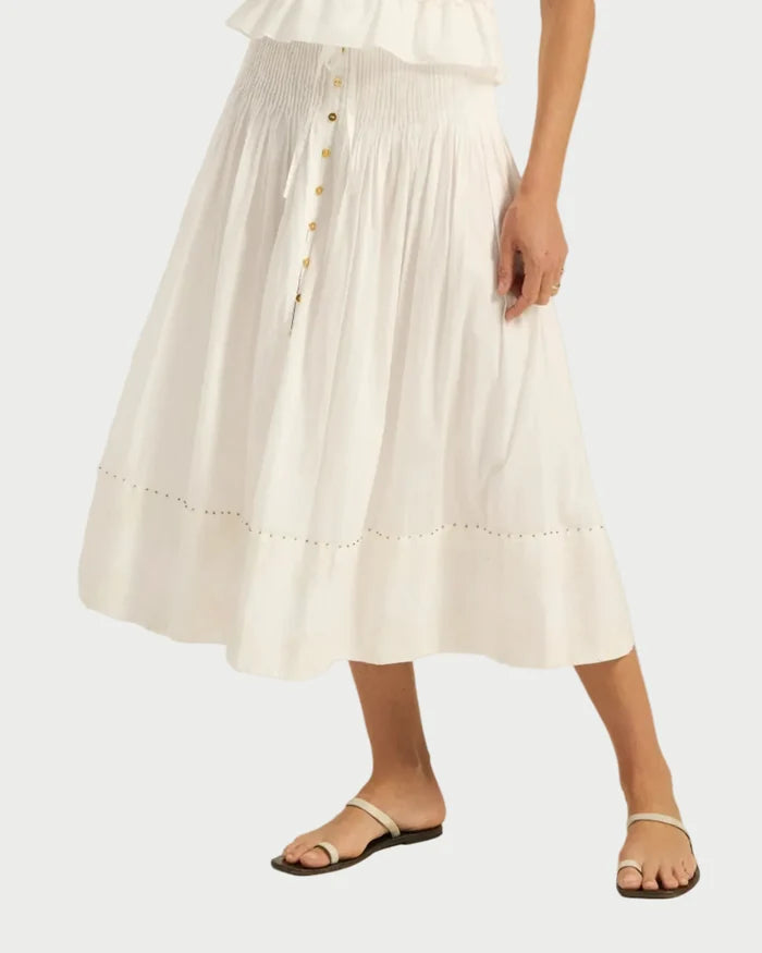 Only the lower half of the body is visible, showing a person wearing the Ilio Nema Diana Skirt—a white voile midi skirt by Ilio Nema with a ruffled waistband and eyelet detail near the hem—paired with flat white sandals.