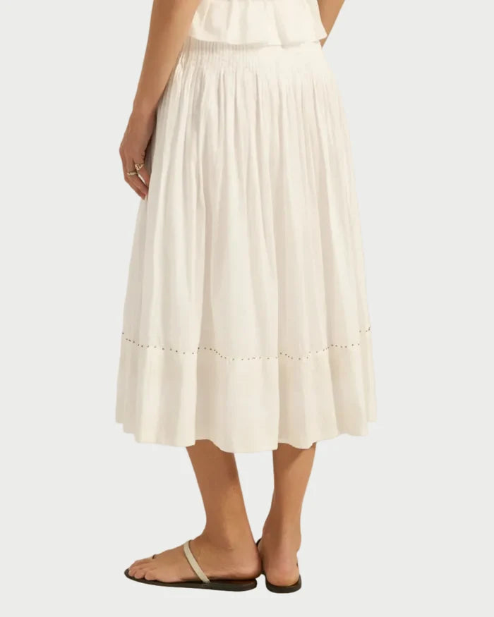 A person stands against a plain light background wearing the Ilio Nema Diana Skirt by Ilio Nema—white voile with a small cut-out near the hem—paired with beige flat sandals and a sleeveless white top.