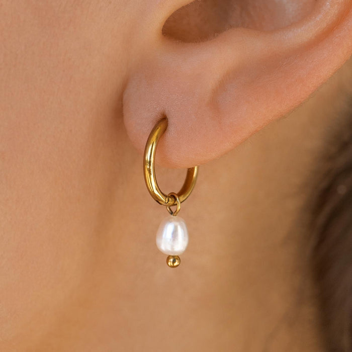 A person wearing a dainty gold huggie earring with a single freshwater pearl dangling from it.
