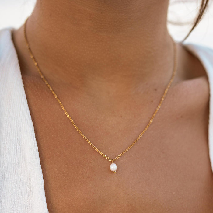 A person wearing a white top showcasing a dainty simple gold necklace with a small freshwater pearl pendant.