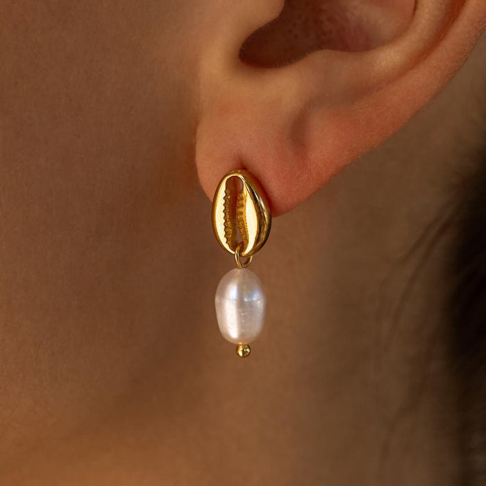 A close-up image of a person wearing a gold puka shell earring with a freshwater pearl drop.